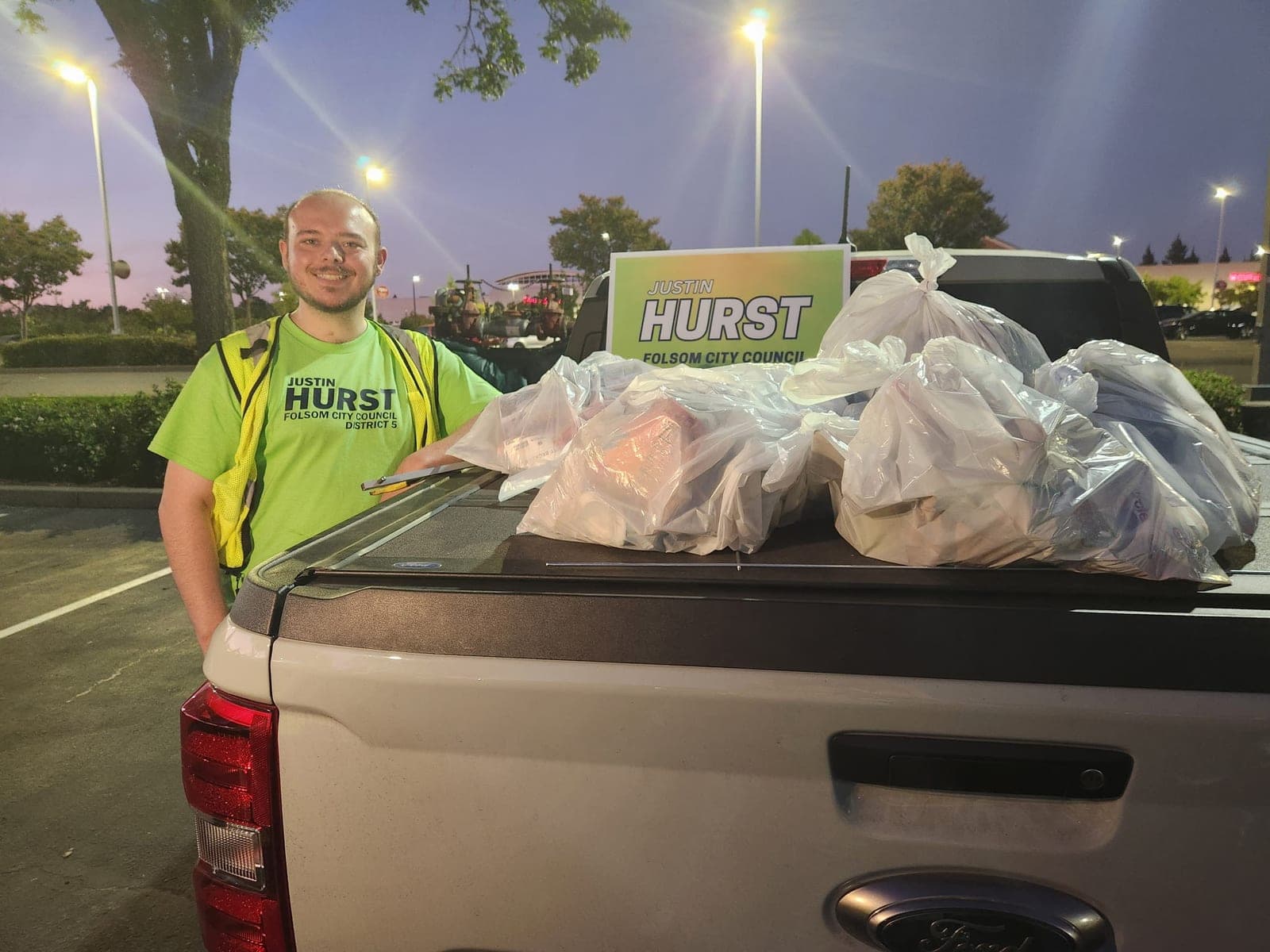 Justin beside a pickup truck bed piled with full trash bags and a Hurst sign at dusk