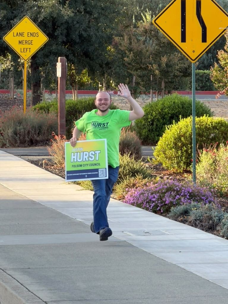 Justin in a green Hurst shirt waving and carrying a Hurst yard sign along a sidewalk